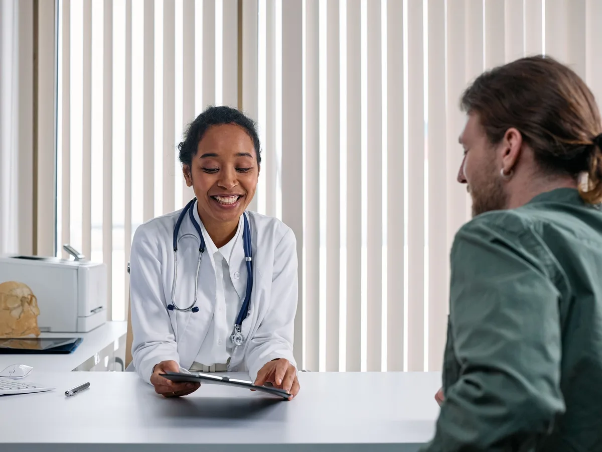 A smiling doctor in a white coat holds a tablet at her desk while talking with a young man in a green shirt.
