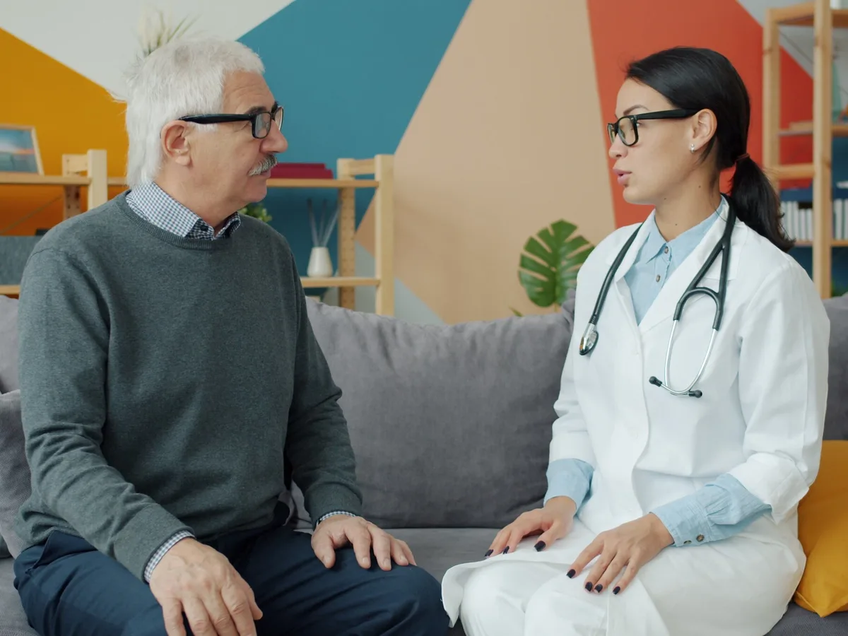 An older man in a gray sweater talks with a doctor in a white coat, both seated on a sofa in a bright room.