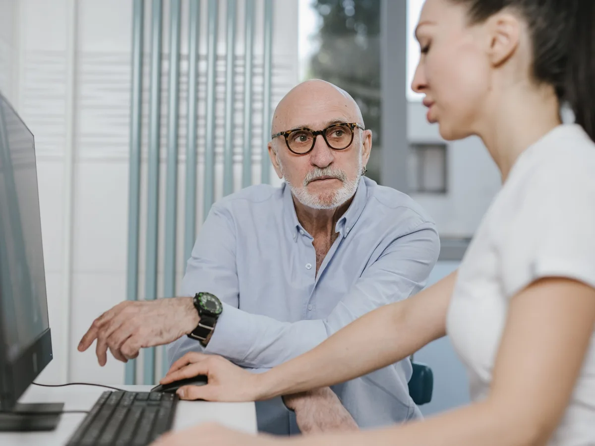 An older bald man with glasses points at a computer screen while a woman beside him types.