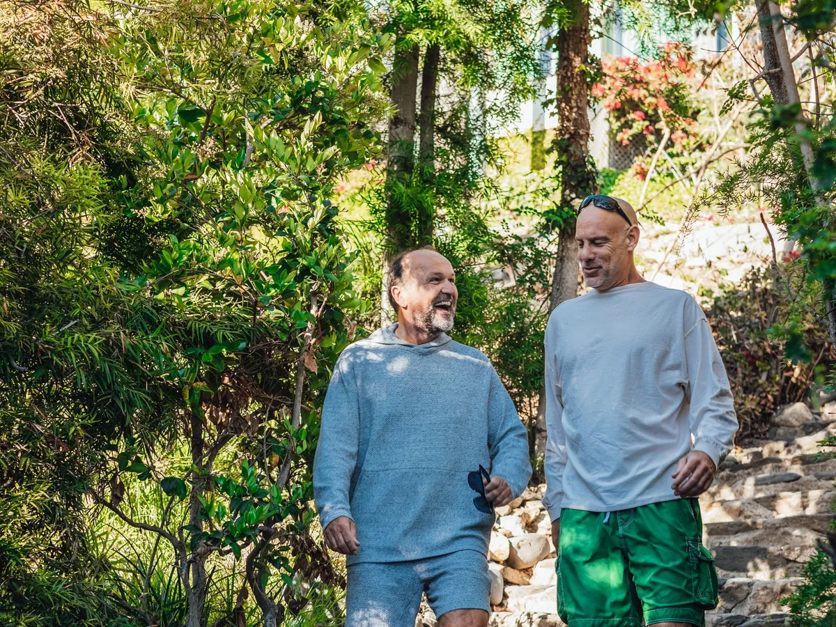 Two men in their fifties laughing as they walk down stone steps on a sunny garden path.